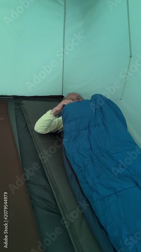 Tourist inside a green camping tent zipping up a warm blue sleeping bag on a mattress.