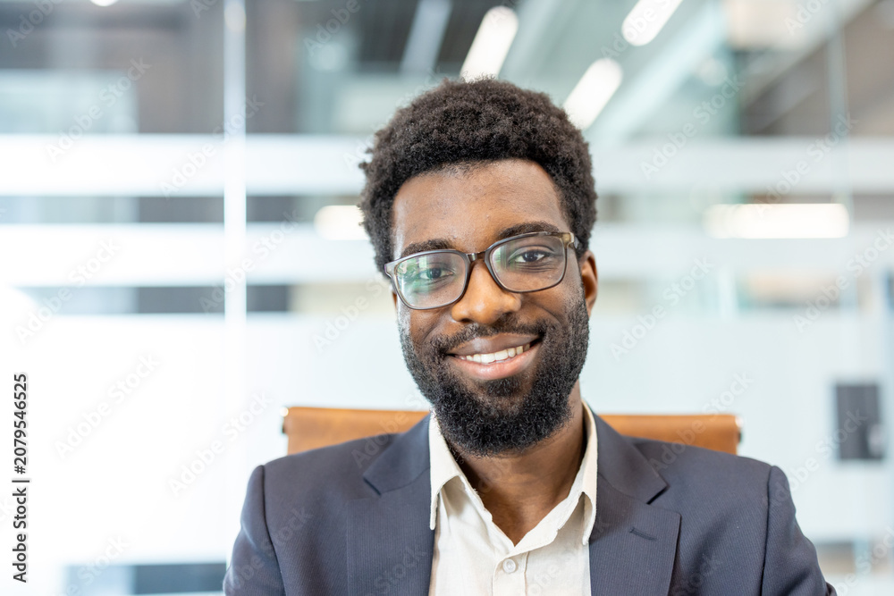Fototapeta premium Young adult professional male wearing eyeglasses and a suit jacket. Smiling confidently while looking at the camera. Portraying success and a friendly business demeanor in a modern office environment