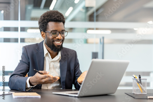 Professional man wearing a suit and glasses. Smiling while engaging in a video call on a laptop. Gesturing and communicating from an office setting. Highlighting online collaboration. Success