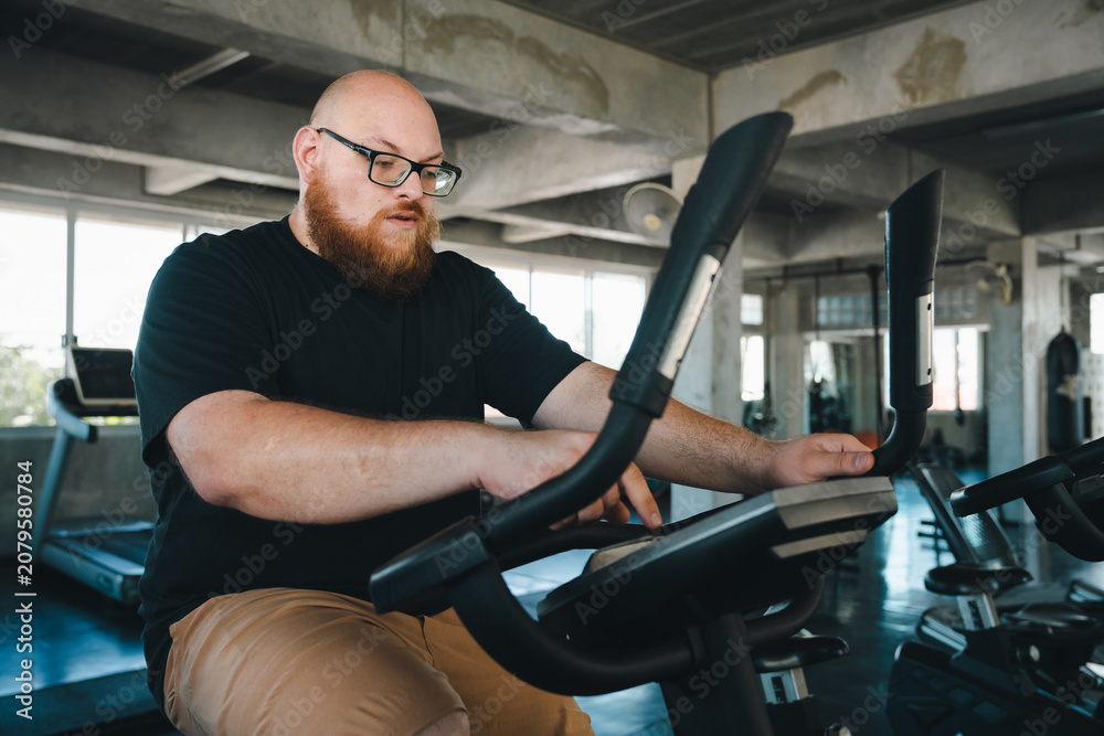 Obraz premium Man exercise bike gym fitness workout indoor bearded eyeglass overweight man using stationary exercise bike in modern gym for cardio training