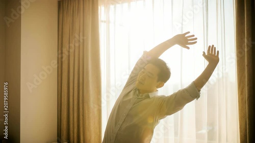 Asian man stretching by a window with bright sunlight. Male professional waking up in a hotel room. Vacation and rest during a business trip