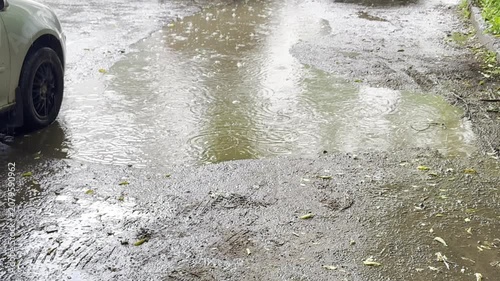 Rainwater accumulates on the muddy ground, creating puddles near parked cars in an urban environment, with visible reflections and ripples as raindrops continue to fall