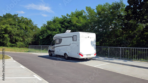 White modern motorhome caravan parked on an asphalt driveway next to a metal railing and green forest trees