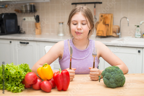 Happy hungry preteen girl holding a fork and knife while sitting at a kitchen table and looking at fresh vegetables. Perfect for healthy eating, nutrition, and family lifestyle concepts.