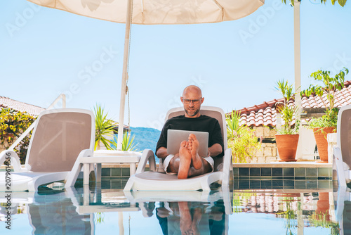 Relaxed man freelancer CEO business owner working laptop on sun lounger by luxury hotel swimming pool. Digital nomad successfully managing company remote tropical vacation sunlit summer day.