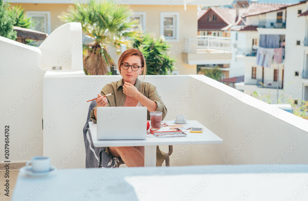 Fototapeta premium Joyful female painter sitting on balcony creating artwork during seaside vacation. Relaxed woman smiling, enjoying creative process and sunny coastal atmosphere, combining leisure, art and inspiration
