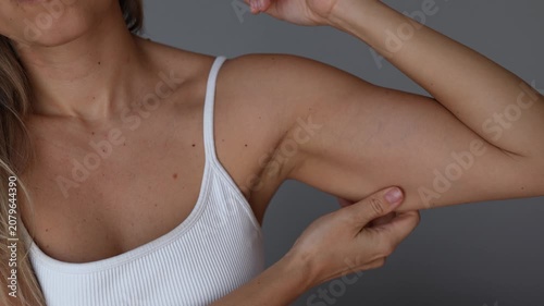 Young woman touches and pinches loose skin on her upper arm, showing body changes after weight loss, sagging skin, soft muscles, excess fat and body insecurity in a wellness concept on grey background