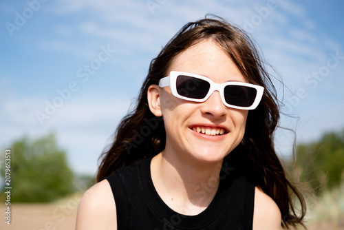 A teenage girl in white sunglasses beams with a joyful carefree smile on a sunlit beach, her hair dancing in the breeze. The teenager radiates pure happiness by the sea on a bright sunny day.