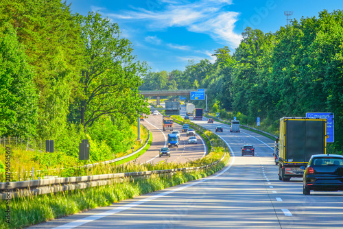 A busy motorway in Germany through a lush green forest with cars and trucks on a sunny day.