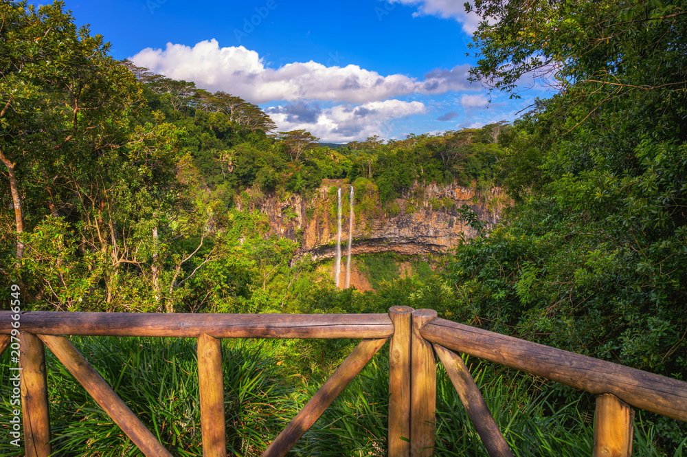 Fototapeta premium View of Chamarel Waterfall framed by a wooden railing and dense tropical forest in southwest Mauritius under a bright sky.