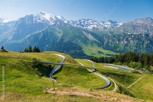 Serpentine paved road curves through alpine meadows with snowy peaks and green ridges near Engelberg, Switzerland.