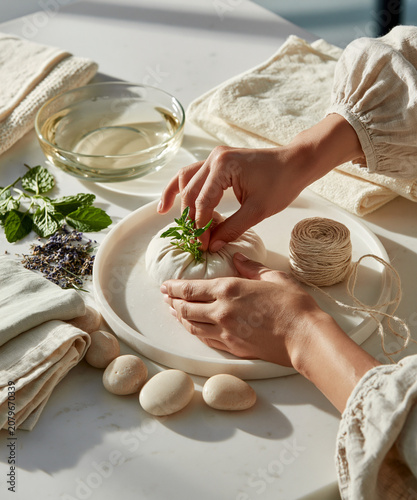 Hands placing fresh herbs on linen spa pouch beside twine and natural wellness items