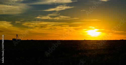 Amarillo, Texas. A serene prairie landscape at a vibrant sunset. The low sun illuminates the sky with brilliant orange hues above the silhouettes of rural buildings and agricultural infrastructure.