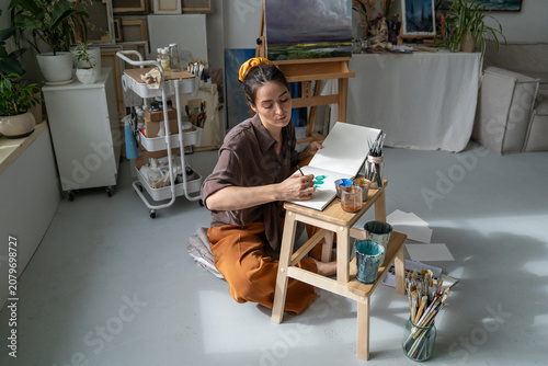 Creative working process at home gallery workshop, thoughtful woman artist making watercolor sketches in album, professional female painter calmly drawing picture ideas in sketchbook using aquarelle.