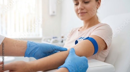 A person sits in a medical office while a healthcare worker ties a band around the arm for a blood draw. The setting is clean and well-lit. The time appears to be daytime