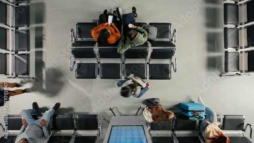 Картината върху платно People on airport chairs talking next to passengers catching plane, hectic passenger movement concept, Travelers with trolleys chatting, waiting to be called to boarding gate, high angle view