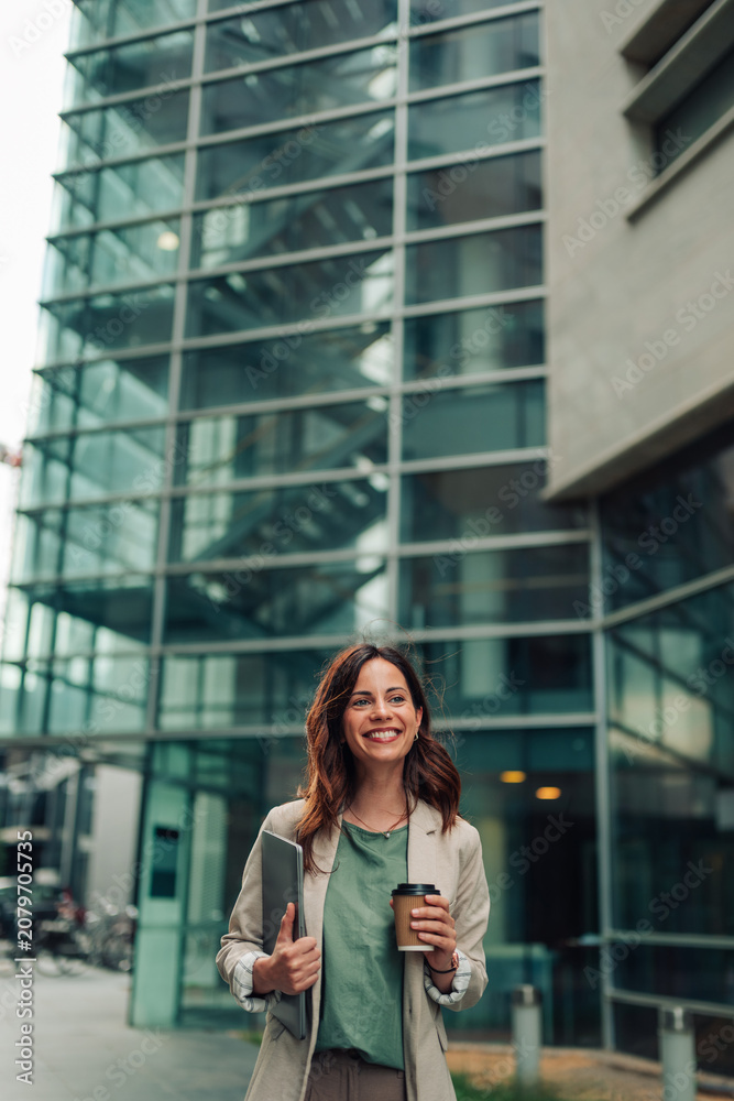 Fototapeta premium Smiling businesswoman walking confidently with a laptop and coffee, preparing for work day in urban environment