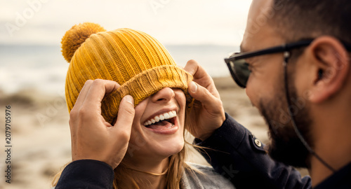 Playful young couple laughing together while man pulls yellow beanie over woman's eyes