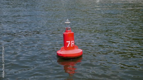 Bright red navigation buoy floats near center on rippled blue green water, showing small lantern top, partial white number marking, soft daylight reflections, and calm maritime