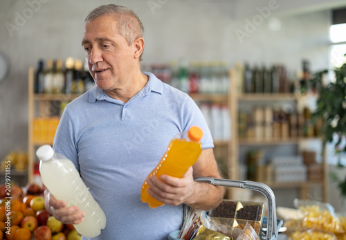 Фотография Concerned old man is deciding between kinds of bottled juice in the supermarket