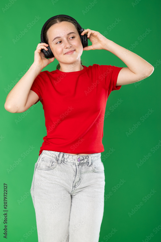 Naklejka premium Passionate teenage girl in red T-shirt and jeans listening and dancing to music on headphones on a green background. Chroma key compositing