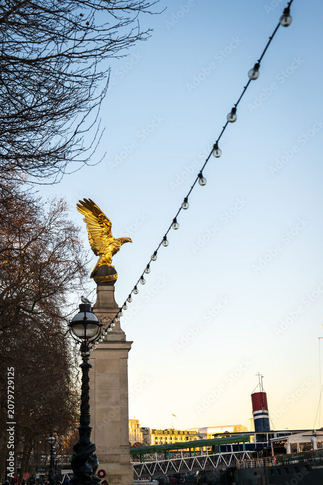 Fototapeta premium Golden Eagle Statue On Riverside Plinth With String Lights And Lamp Post At Dusk In London