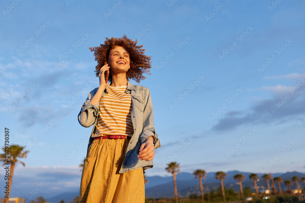 Fototapeta premium Cheerful young woman strolls outdoors talking on a mobile phone under a clear blue sky with palm trees, wearing a striped shirt, denim jacket, and mustard pants.