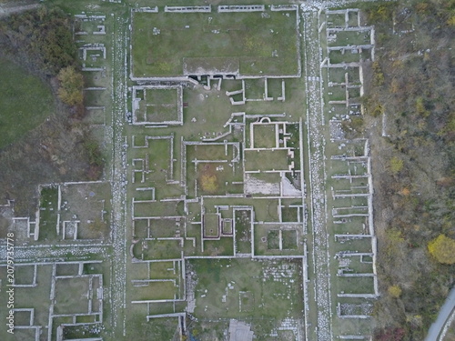 Aerial view of the ancient Roman archaeological site of Alba Fucens with stone foundations, walls, and pathways surrounded by trees in Albe, Abruzzo, Italy.