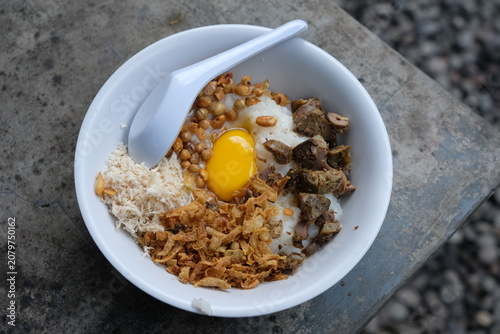 Bubur ayam. Chicken porridge topped with shredded chicken, fried soybeans, raw egg yolk, fried shallots, and minced chicken liver and gizzard. Served in a white ceramic bowl. Indonesian food.