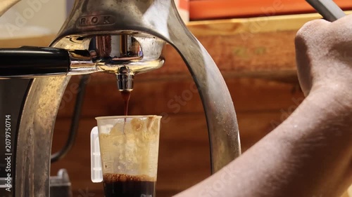 Close up of a manual lever espresso machine brewing a fresh shot of coffee with thick crema layer into a clear plastic measuring cup.