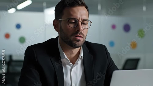 Stressed businessman working on laptop in modern office, feeling overwhelmed and tired.