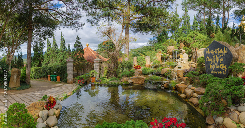 Konstfotografi DALAT, VIETNAM, 5 November 2025 – A serene garden of Thien Vien Truc Lam features a tranquil pond, a cascading waterfall with a Buddha statue, and a large stone inscribed with Vietnamese calligraphy