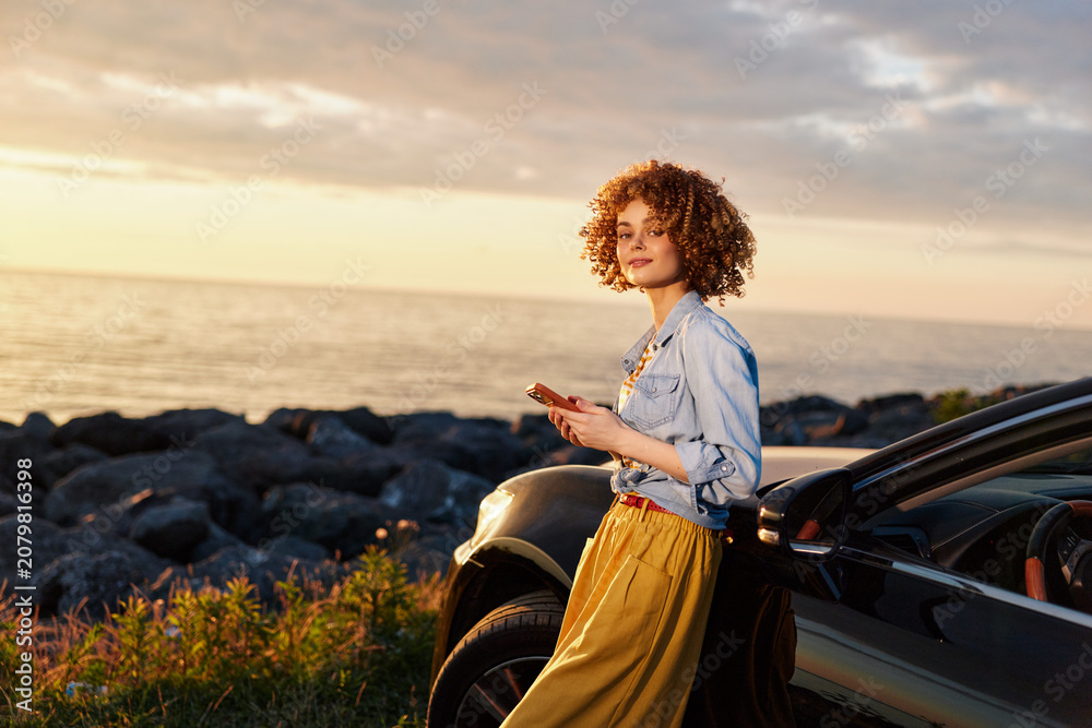 Fototapeta premium A young woman with curly hair stands beside a parked car by the rocky coast at sunset, exuding calm confidence and a relaxed, aspirational mood.