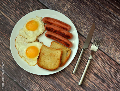 A hearty breakfast: two fried eggs with sausages and toasted bread in a round bowl on a wooden table, with cutlery nearby.