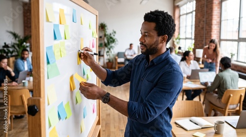 Man writing on sticky notes on a whiteboard in a modern office with colleagues collaborating in the background