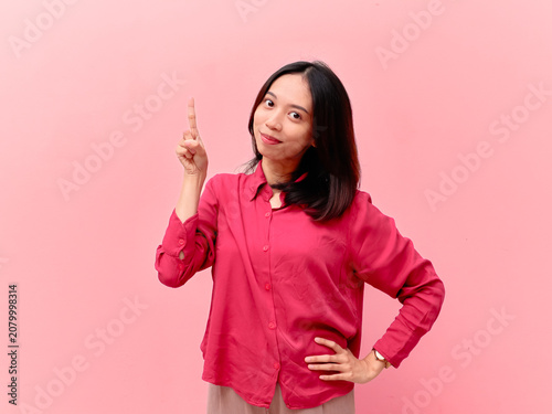 Smiling Asian woman pointing up with one finger, standing against solid pink background with copyspace.