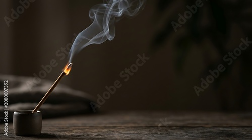 Burning incense stick with wispy smoke on dark wooden surface
