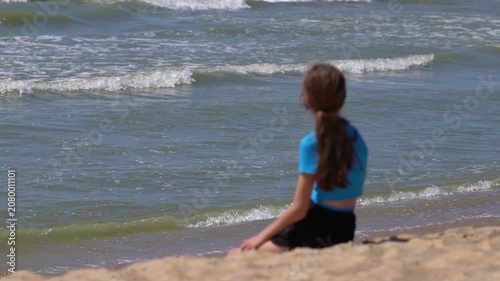 Young girl with long braided hair sitting alone on sandy beach surrounded by grass looking out over calm sea under cloudy sky