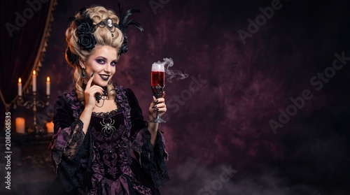 Glamorous woman in dark purple gothic baroque vampire costume with black roses in wig, dramatic makeup, holding glass of smoky red cocktail against moody dark background.