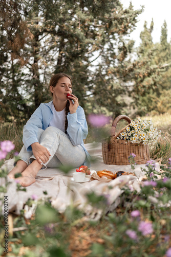 Picnic Flowers Woman: Relaxing woman eats strawberry during daytime picnic with flower basket in forest.
