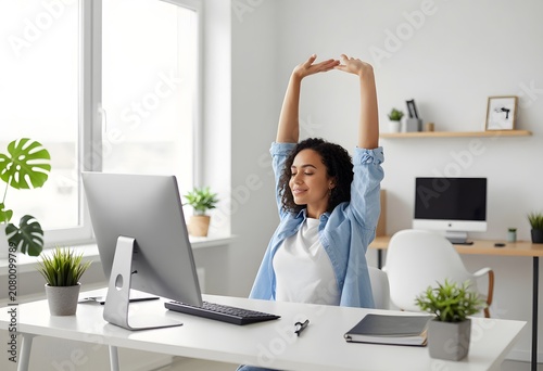 Young woman stretches her arms above her head at a clean desk with a computer monitor, taking a refreshing break from work in a bright home office environment.
