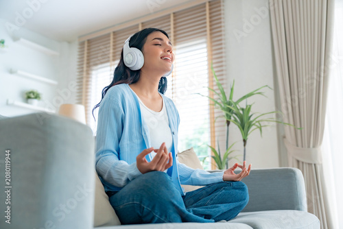 Happy young Asian woman practicing yoga and meditation at home sitting on couch in living room in lotus position and relaxing with closed eyes. Mindful meditation and wellbeing concept