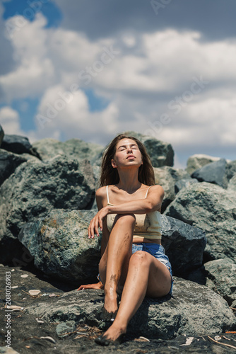 Young woman sits on rocks at the beach under a blue sky with clouds during daytime and enjoys the warm sunlight on her face
