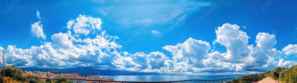 Fototapeta premium Panoramic coastal town and calm blue sea under towering white cumulus clouds, terracotta rooftops and green vegetation along a sunlit shore, serene and expansive
