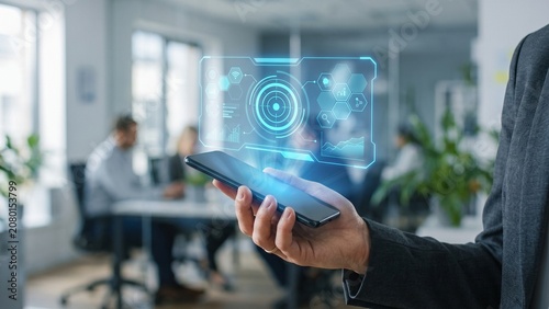 Businessman Hand Holding Smartphone with Futuristic Holographic Data Interface in Modern Office