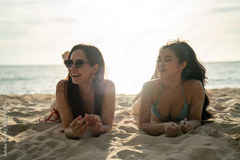 Fototapeta premium Two happy young women in colorful bikinis lying on a beautiful tropical sandy beach, smiling and talking. Friends enjoying a relaxing summer vacation, sunbathing and tanning by the sea.