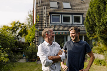Smiling father with arms crossed looking at cheerful son while standing in backyard