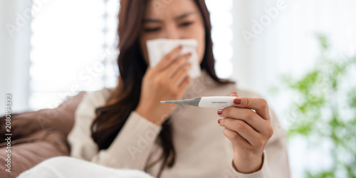 Fotografía A woman holding fever thermometer suffering from a cold, the flu, or allergies is blowing her nose with a tissue while lying on a sofa in the living room