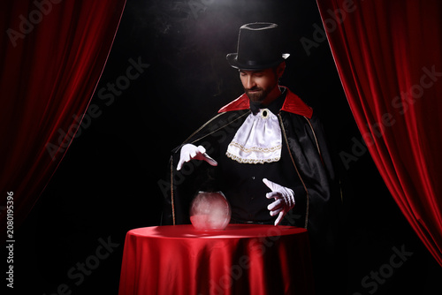 Fotografie Male magician with crystal ball at table near red curtains on dark background