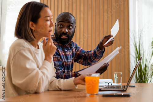 A man examines paperwork closely as his partner reacts with concern. Couple managing household finances.